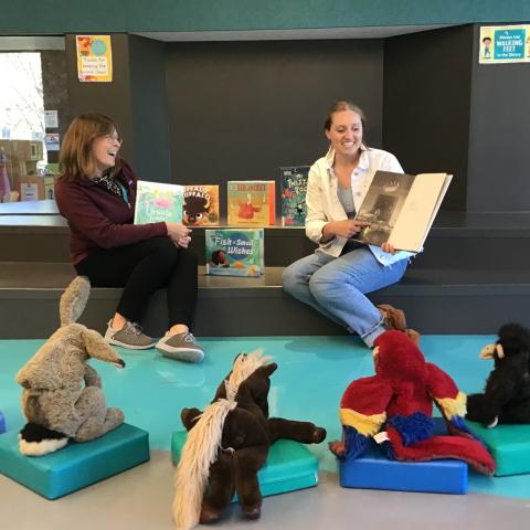 Stuffed animals sitting in a semi-circle around two women reading picture books.