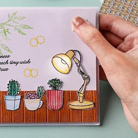 Photo of a woman's hand holding a greeting card with a picture of an illustrated lamp on it. The lamp is lit by a real tiny light.