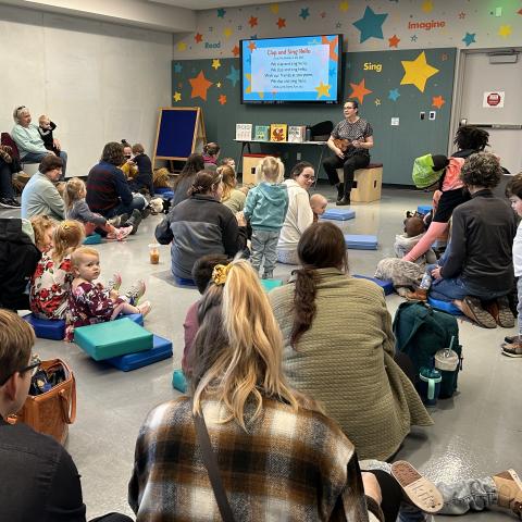 A group of families sitting and watching a woman play a ukulele. 