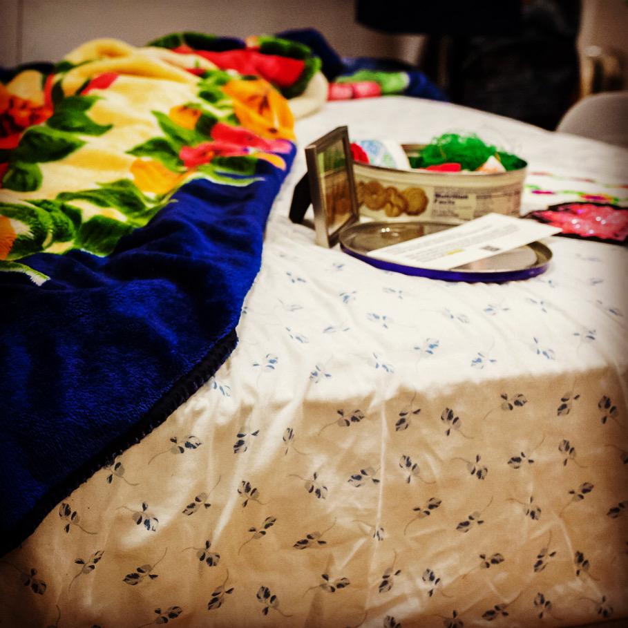 From the exhibit, a collection of objects on a bed, including a flower print blanket, a picture frame, and a cookie tin.