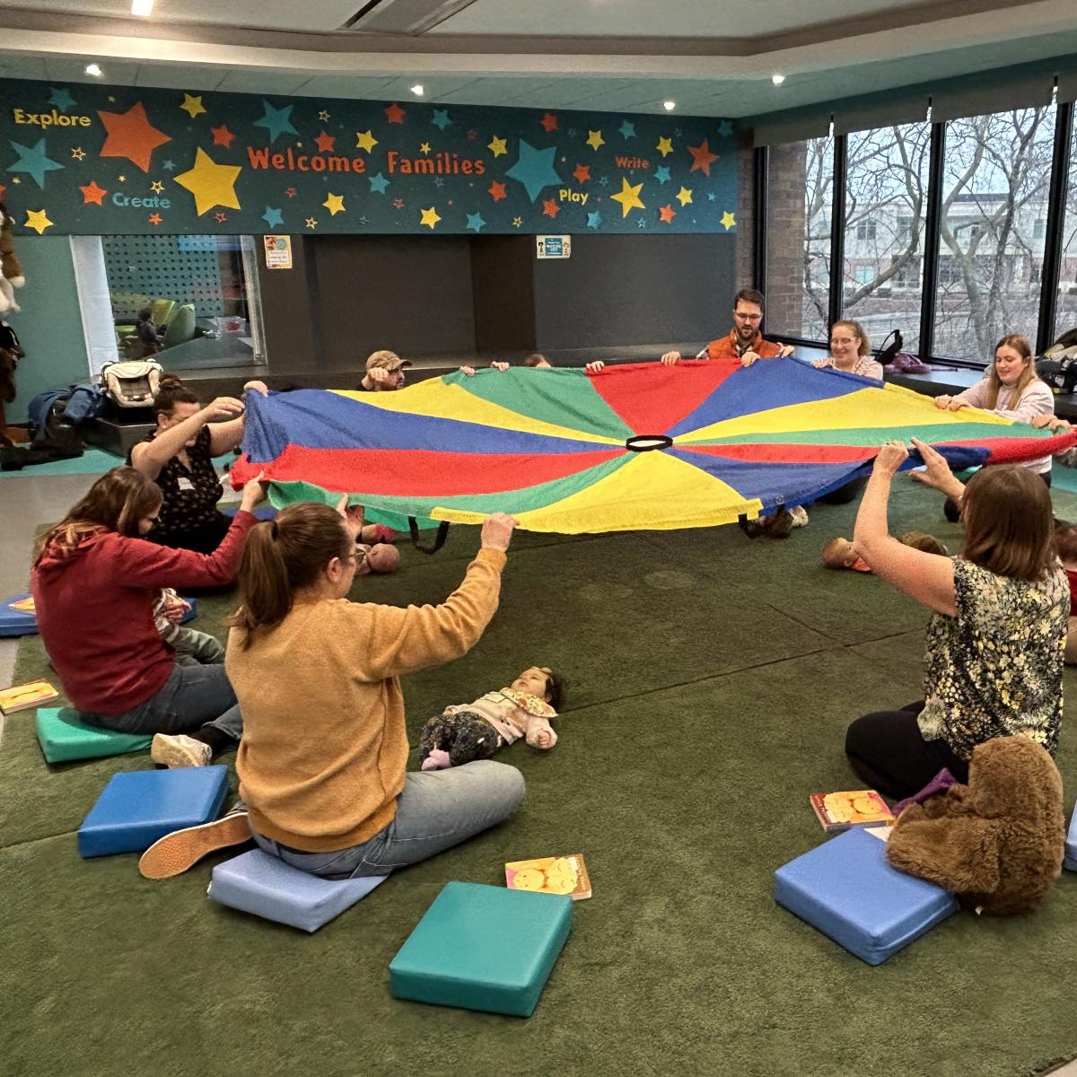 Adults sitting in a circle, holding a colorful parachute in the air above little babies.