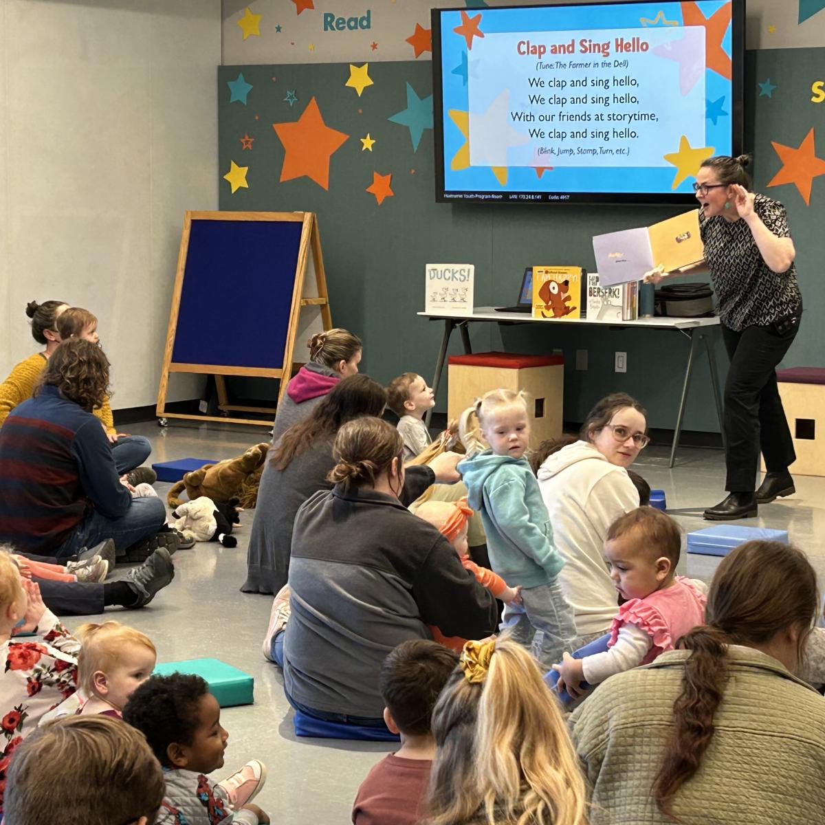 A group of kids and adults sitting on the floor, watching a female presenter reading a book. 